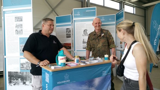 Kleiner Infostand mit zwei Männern, die mit einer Besucherin sprechen.