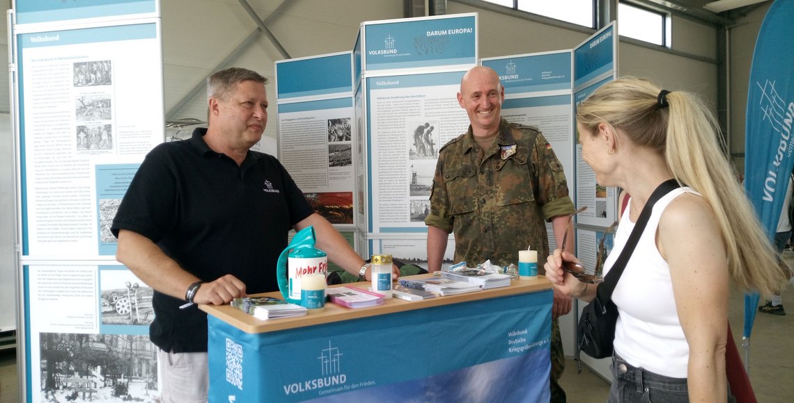 Kleiner Infostand mit zwei Männern, die mit einer Besucherin sprechen.