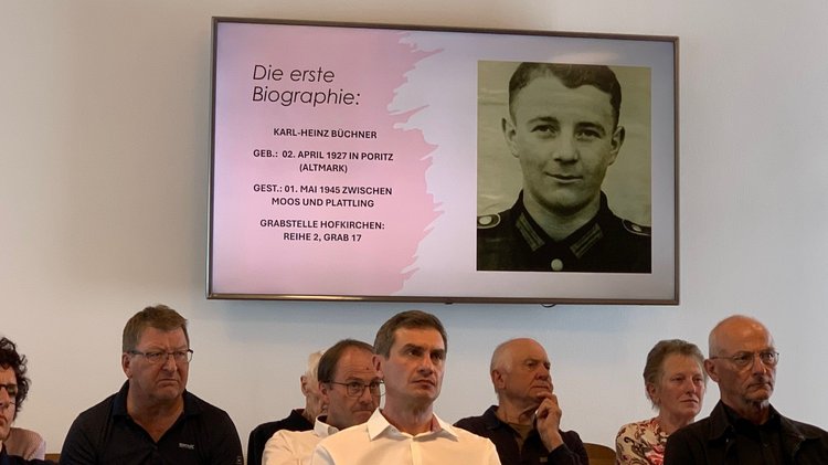 Personen, die einem Vortrag zuhören. Im Hintergrund eine Bildschirmprojektion: Vorstellung der Zwischenergebnisse des Biographieprojekts zur Kriegsgräberstätte Hofkirchen